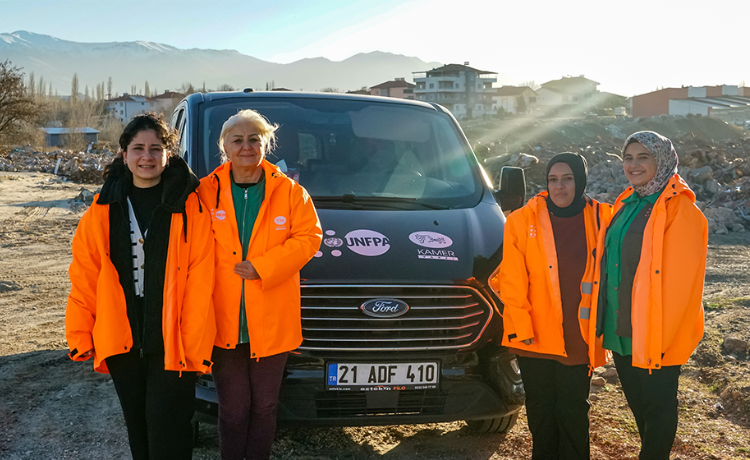 Four women in bright orange jackets stand in front of a car amid rubble caused by the 2023 earthquake in Türkiye.