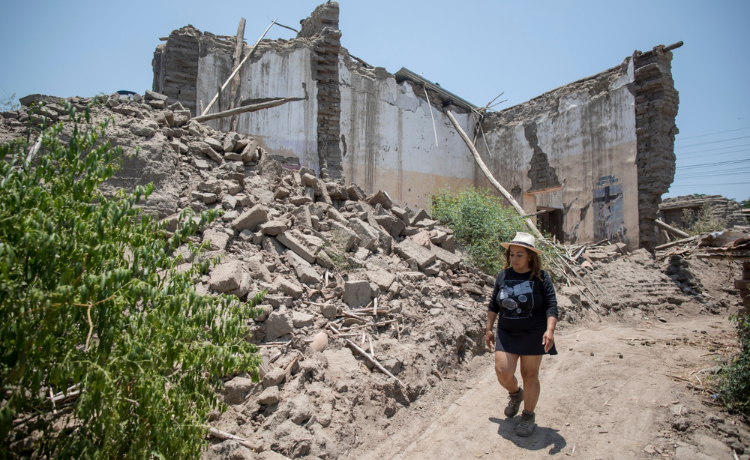 A woman walks the dirt path next to a destroyed building.