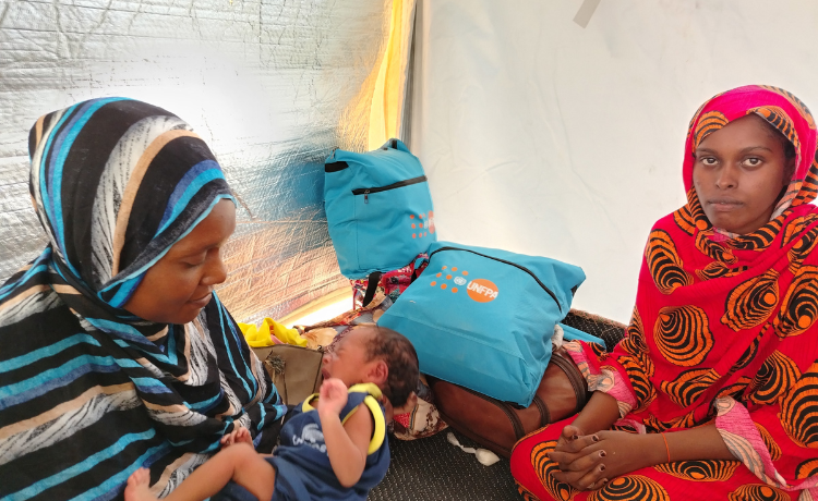 Una mujer vestida de rojo sonríe ligeramente a la cámara sentada en una tienda de campaña, con bolsas del UNFPA visibles detrás de ella. Otra mujer en rayas azules y negras sostiene a un bebé que llora.]