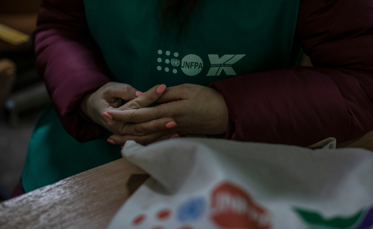 A woman wearing orange nail polish clasps her hands in her lap as she sits at a desk, she is wearing a green top with UNFPA visible on it