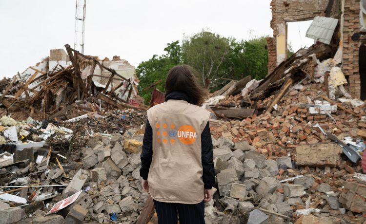 A woman stands in front of destroyed houses with only rubble left; she has her back to the camera and is wearing a UNFPA vest