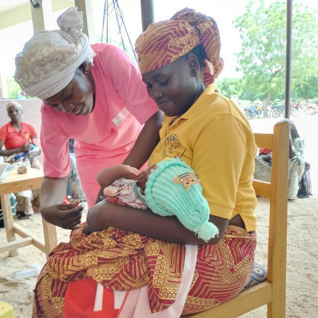A midwife bends down to assist a woman seated holding a baby.