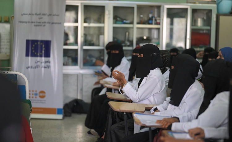 A group of women in medical coats sit behind desks looking towards the front of the classroom