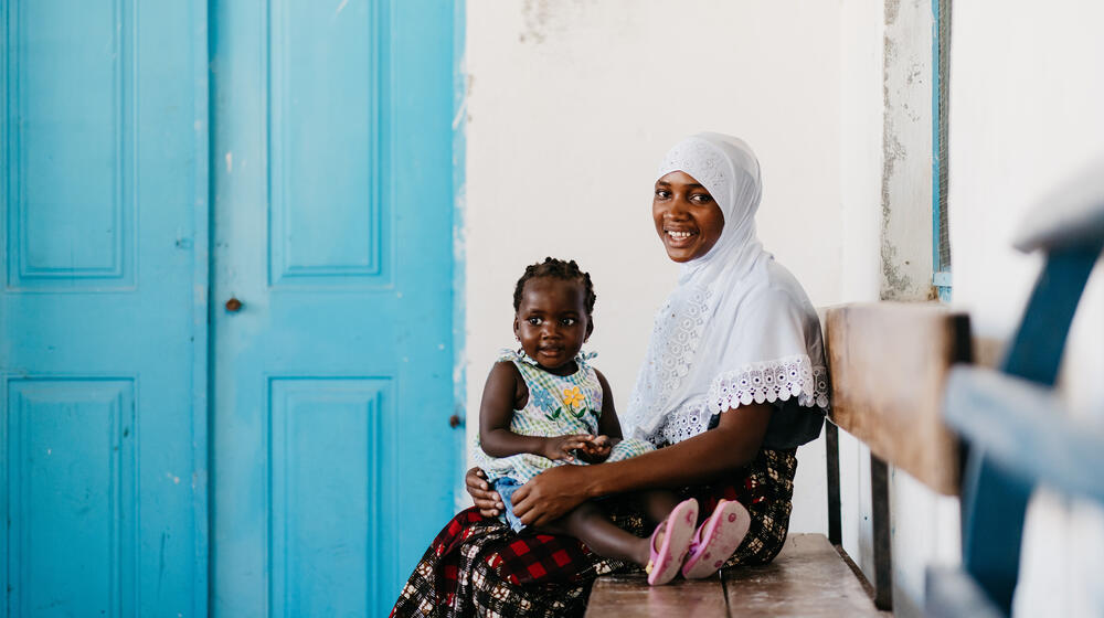 A woman sits on a bench and holds her young daughter on her lap. 