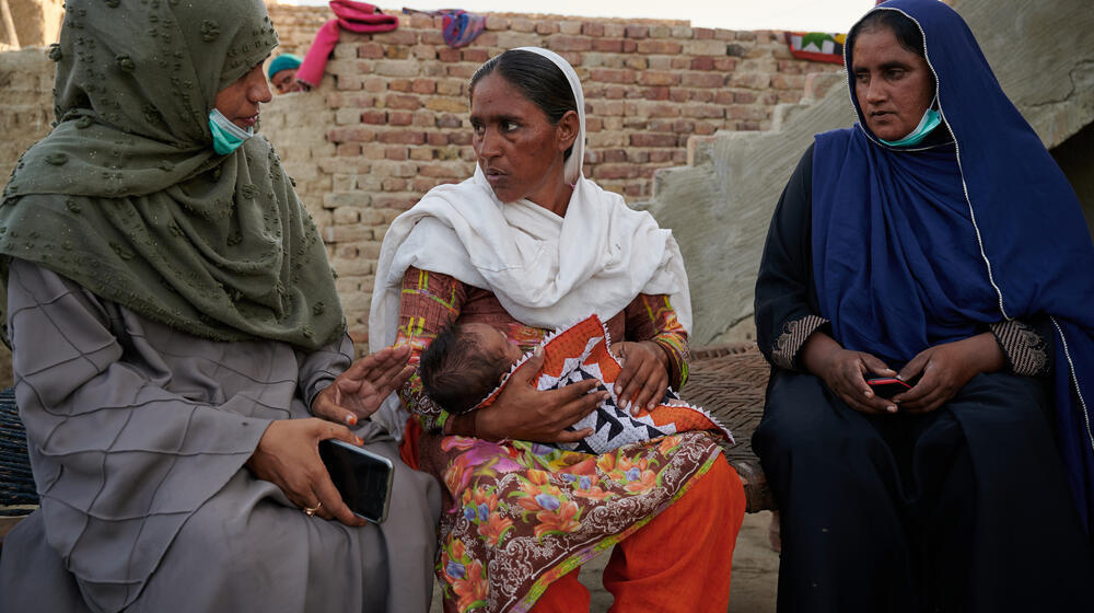 Three women are speaking and seated together at the foot of a brick building. One of the women is holding a young infant in her arms.