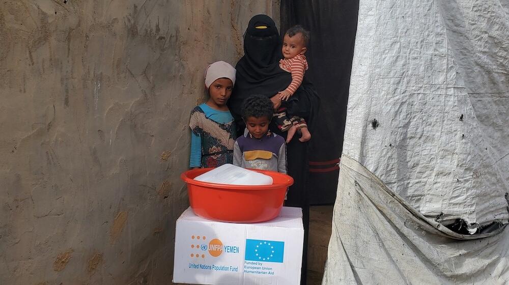 Une femme se tient avec ses trois enfants devant un carton et une bassine.