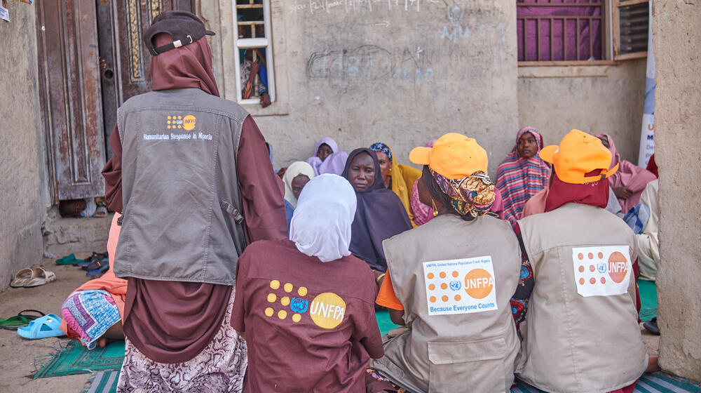 Mujeres con el logotipo del UNFPA bordado en la ropa hablan con un grupo de mujeres sentadas en mantas en un patio.