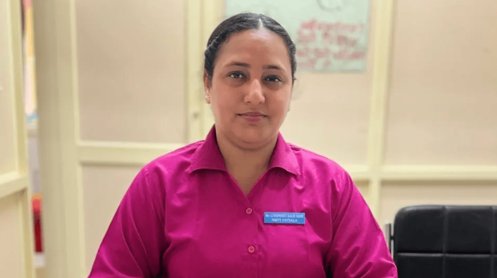 Una mujer con uniforme rosado de enfermera sentada en un escritorio frente a un libro y un celular.