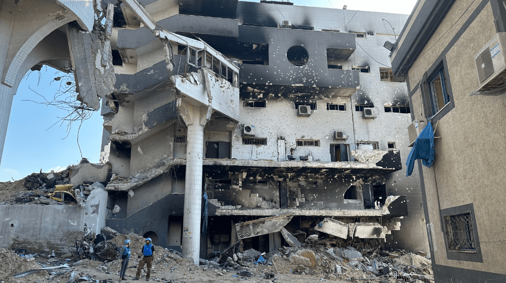 Two United Nations personnel in blue helmets stand in the ruins of a hospital