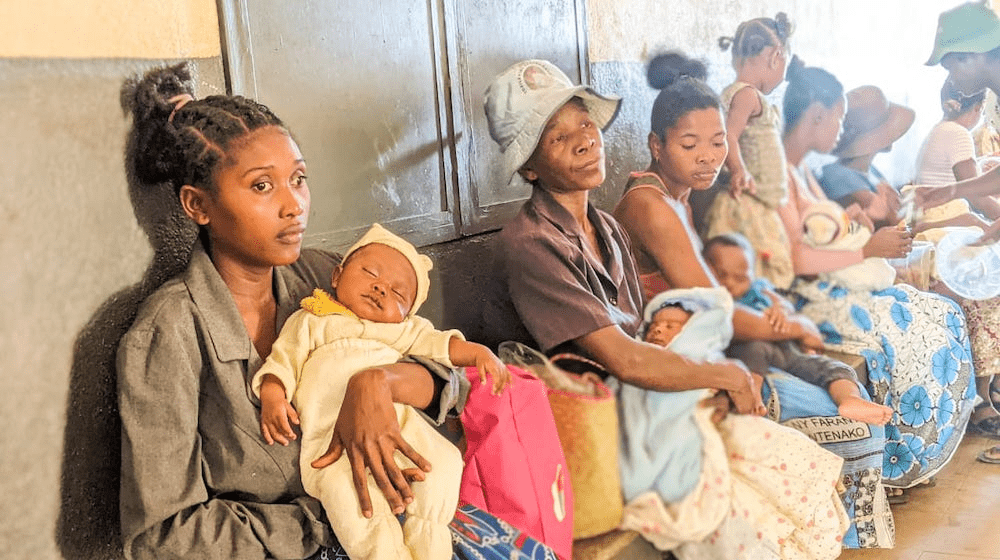 A young woman sits against a wall holding a newborn baby at the head of a line of other women and children.