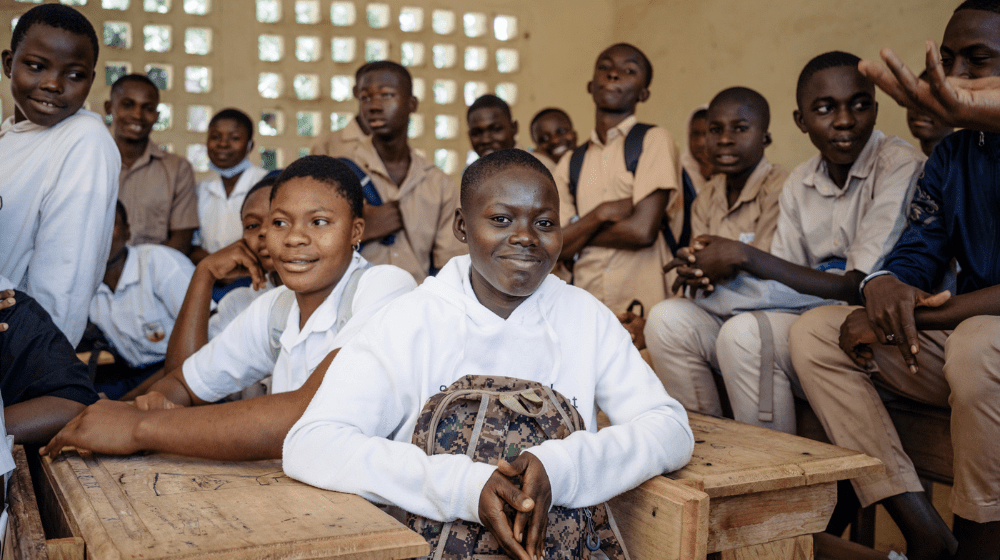 A group of students sit around and on top of desks in a classroom