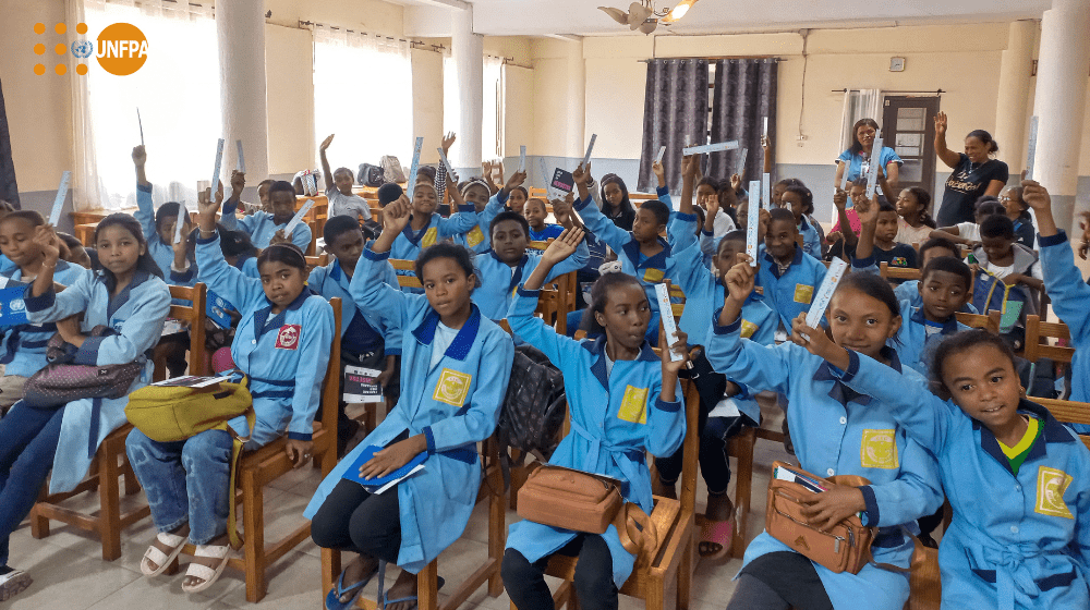 A group of young students in blue uniforms raise their hands while seated in a classroom