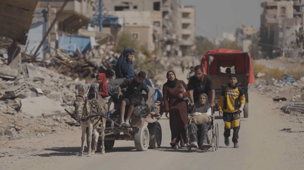 A group of people, one in a wheelchair and several on a donkey cart, travel along a dusty road surrounded by destroyed buildings and rubble