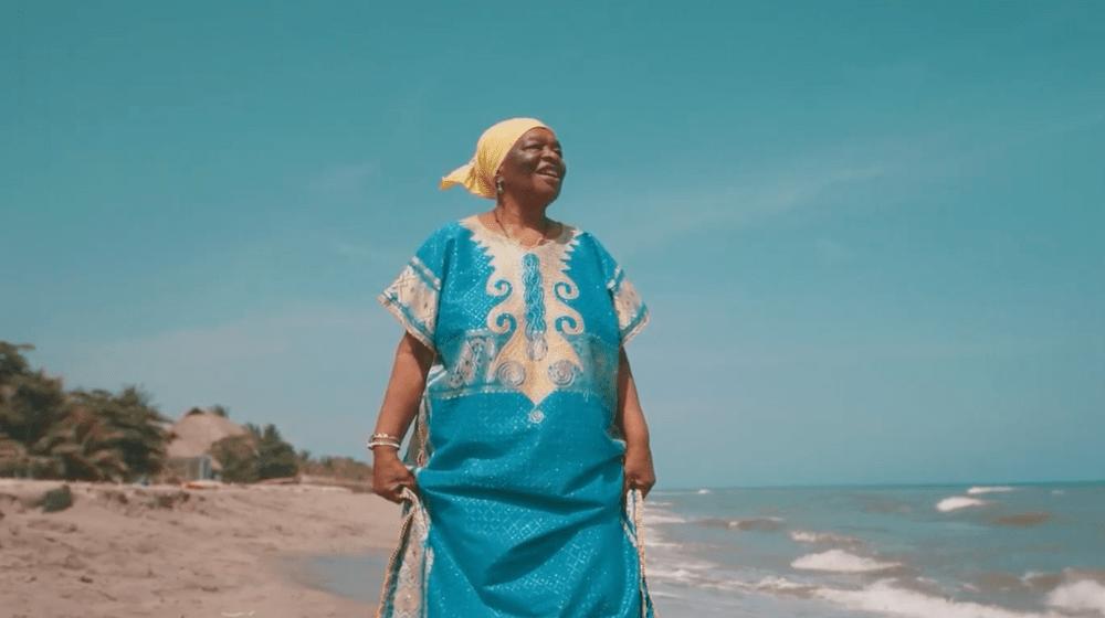 An older Afrodescendent woman in a blue dress walks along the beach.