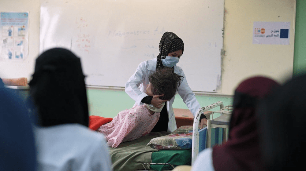 A woman in a dark headscarf and white medical coat holds a dummy of a female patient on a hospital bed