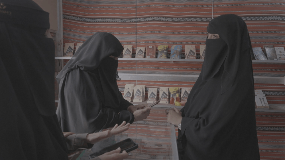  Tres mujeres con niqabs negros y abayas hablan entre sí en una habitación pequeña, dos de ellas con teléfonos celulares.