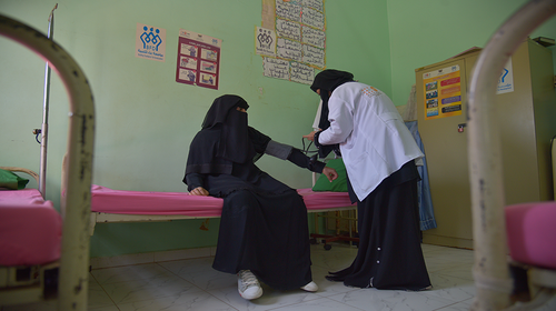 A doctor in a white coat checks the blood pressure of a woman sitting upright on a bed in a black niqab