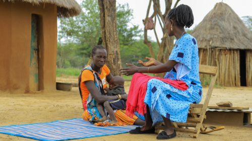  Josephine Telo, habillée en orange, est assise sur un tapis dans une cour. Assise sur une chaise, Consolata Aleper porte une robe bleue.