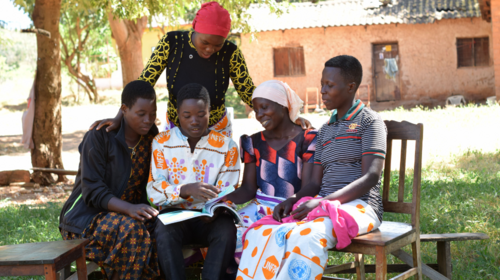 Des jeunes sont rassemblé·e·s à l’ombre dans une cour, et regardent un livre ensemble. L’un d’entre eux porte un t-shirt orné du logo de l’UNFPA. Trois autres sont vêtus d’une jupe portefeuille portant aussi le logo de l’UNFPA.