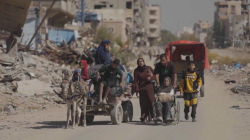 A group of people, one in a wheelchair and several on a donkey cart, travel along a dusty road surrounded by destroyed buildings and rubble