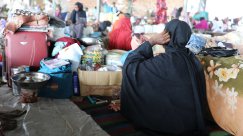  Several women sit with their belongings in an open-air temporary shelter.